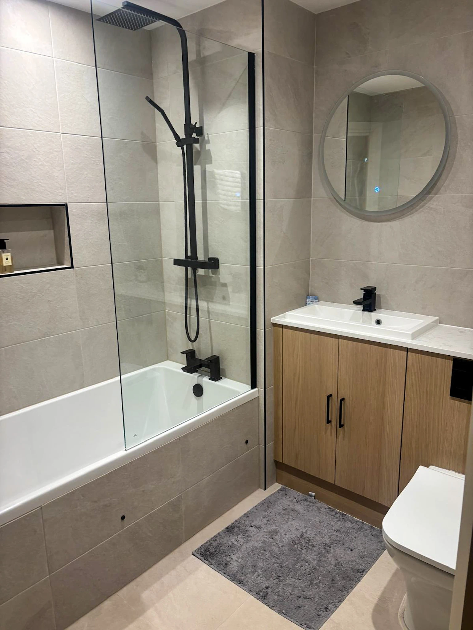 Modern bathroom featuring a glass shower, bathtub, circular mirror, wooden cabinetry, and gray tiles.